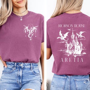 May include: A mauve t-shirt with a white dragon and star design on the front and a castle with dragons on the back. The text "RIORSON HOUSE ARETIA" is printed on the back of the shirt.