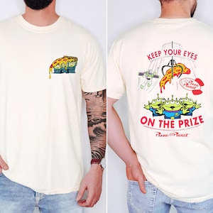 May include: Cream-colored t-shirt featuring a Pizza Planet design. The front displays a pizza slice with aliens, while the back showcases the text "KEEP YOUR EYES ON THE PRIZE" with a claw machine, Buzz Lightyear, and more aliens.