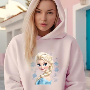 May include: A pink hoodie with a graphic of a cartoon princess with blonde hair and a blue dress. The princess is surrounded by snowflakes.