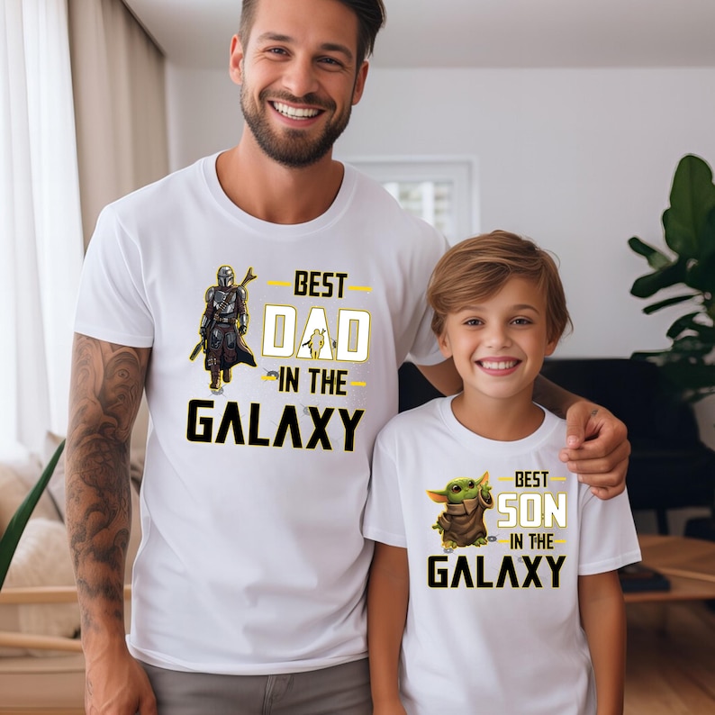 Pu&ograve; includere: Magliette bianche con grafica abbinata. Una maglietta dice "BEST DAD IN THE GALAXY" con una grafica di Mandaloriano. L'altra dice "BEST SON IN THE GALAXY" con una grafica di Grogu. Le magliette sono indossate da un padre e un figlio.