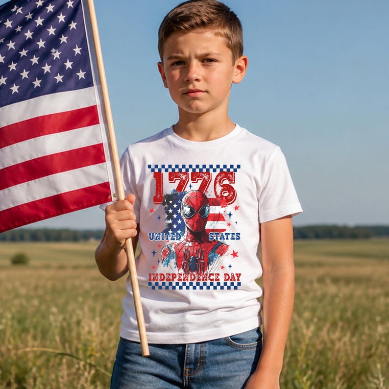 May include: A white t-shirt with a graphic of Spider-Man, the American flag, and the text "1776 United States Independence Day." The boy is holding an American flag. The background is a field of grass and a blue sky.