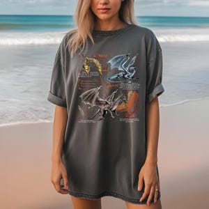 Peut inclure: T-shirt gris fonc&eacute; avec quatre illustrations de dragons et du texte. Les dragons sont repr&eacute;sent&eacute;s en or, bleu et gris, dans diff&eacute;rentes poses. Le t-shirt a des manches courtes et une coupe d&eacute;contract&eacute;e.