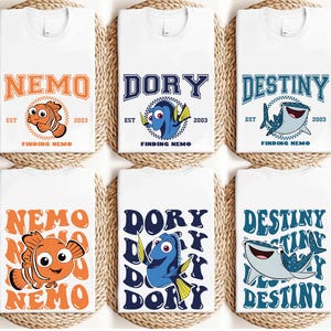 May include: White t-shirts featuring designs from the movie "Finding Nemo." The shirts display the names "Nemo," "Dory," and "Destiny" in various fonts and colors, along with character illustrations and the text "EST 2003." The shirts are arranged on a woven mat.