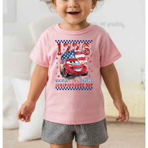 A young girl wearing a pink t-shirt with a graphic of a red car and an American flag, smiling at the camera.