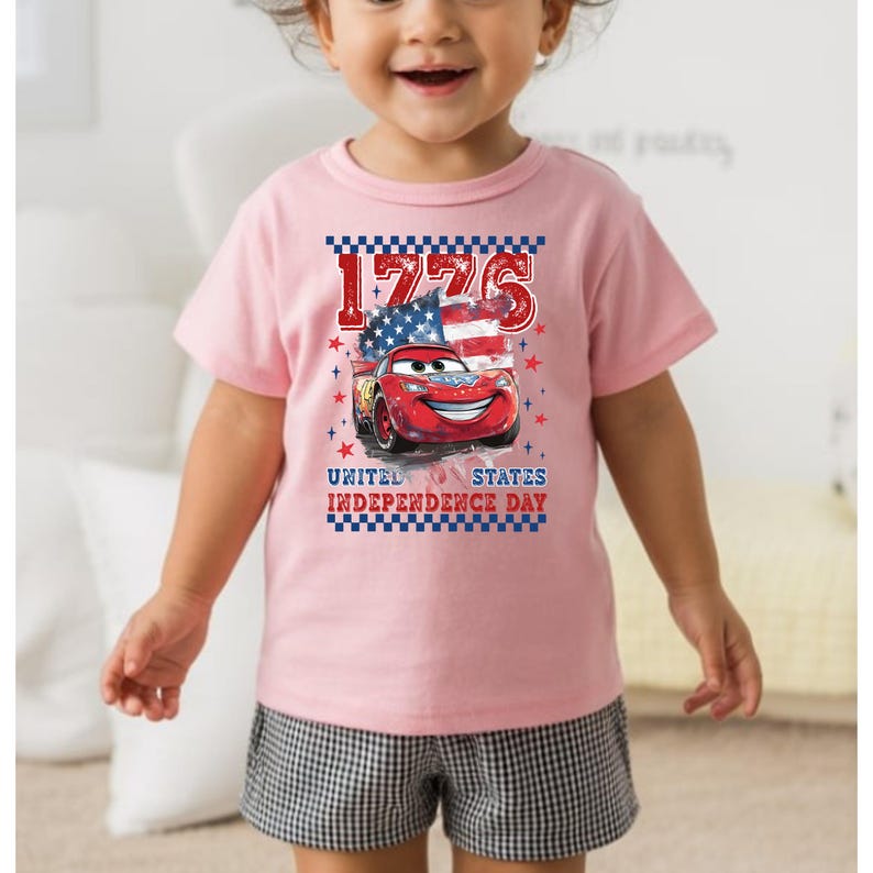A young girl wearing a pink t-shirt with a graphic of a red car and an American flag, smiling at the camera.