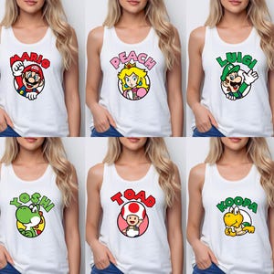 May include: A collection of six white tank tops, each featuring a circular graphic of a Nintendo character. The designs include Mario, Peach, Luigi, Yoshi, Toad, and Koopa, with their respective names printed in bold, colorful letters above each character.