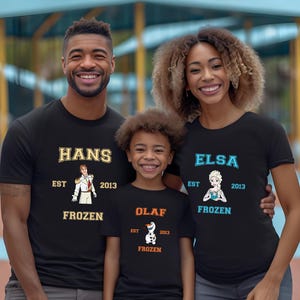 May include: A family of three wearing black t-shirts with Frozen character graphics. The shirts have the text "Hans Est 2013 Frozen", "Elsa Est 2013 Frozen", and "Olaf Est 2013 Frozen".