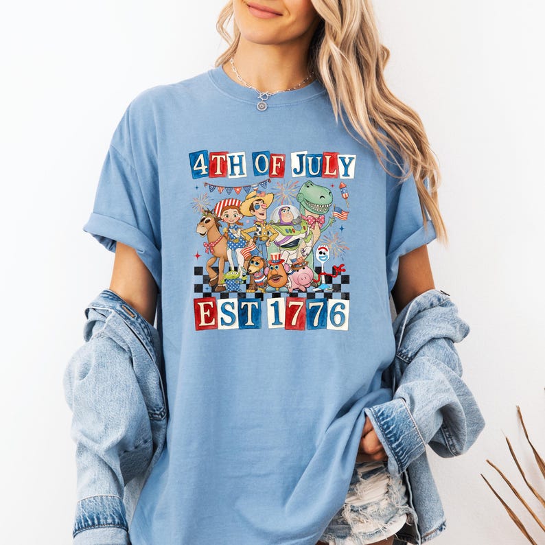 A woman wearing a blue t-shirt with a graphic design depicting cartoon characters and text related to the 4th of July holiday.