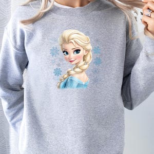 May include: Gray sweatshirt with a printed image of Elsa from Frozen. Elsa is wearing a blue dress and has long blonde hair. There are snowflakes around her.