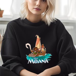 May include: Black sweatshirt featuring a colorful graphic of characters in a boat with a sail. The design includes the text "Disney Moana" in white and blue. The sweatshirt is a crew neck style.