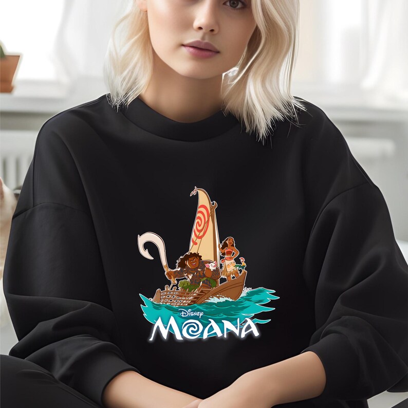 May include: Black sweatshirt featuring a colorful graphic of characters in a boat with a sail. The design includes the text "Disney Moana" in white and blue. The sweatshirt is a crew neck style.