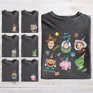 Puede incluir: Camisetas gris oscuro con varios personajes de Toy Story, incluyendo a Woody, Buzz Lightyear y Jessie. Cada camiseta tiene un gr&aacute;fico de personaje y la palabra "Name" debajo. La camiseta m&aacute;s grande muestra varios personajes.