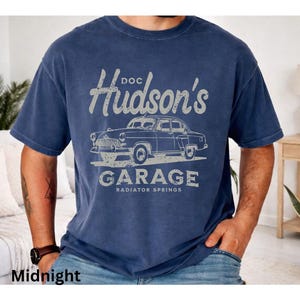 Op de afbeelding: Nachtblau T-shirt met een vintage auto-afbeelding en de tekst "Doc Hudson's Garage, Radiator Springs". Het ontwerp heeft een distressed, vintage esthetiek. Het shirt is geschikt voor casual wear.
