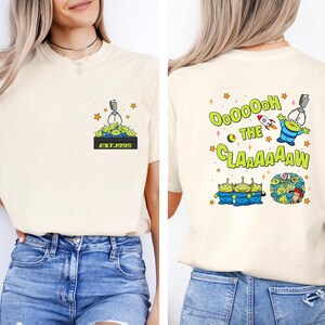 May include: Cream-colored t-shirt featuring a cartoon design of green aliens and a claw machine. The front has a small graphic with the text "EST.1995". The back displays the phrase "Ooooooh the Claaaaaw" with more alien graphics.