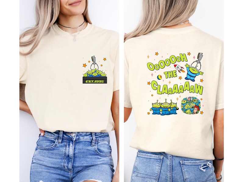 May include: Cream-colored t-shirt featuring a cartoon design of green aliens and a claw machine. The front has a small graphic with the text "EST.1995". The back displays the phrase "Ooooooh the Claaaaaw" with more alien graphics.