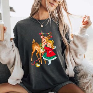 Op de afbeelding: Donkergrijze sweatshirt met een vintage kerstafbeelding. De afbeelding toont een hert met kleurrijke lichtjes op zijn gewei, een meisje in een rode jas en een klein mandje. Het ontwerp heeft een retro-uitstraling, perfect voor de feestdagen.