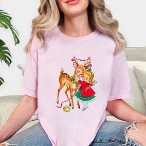 Op de afbeelding: Lichtroze T-shirt met een vintage kerstillustratie. De afbeelding toont een hert versierd met kerstverlichting en een meisje in een rode jas en groene jurk. Het hert en het meisje omarmen elkaar, wat een feestelijke kerstsc&egrave;ne cre&euml;ert.