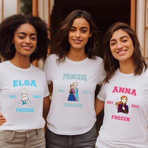 May include: Three white t-shirts with Frozen characters and text. The first shirt says "Elsa Est 2013 Frozen" and features Elsa. The second shirt says "Princess Est 2013 Frozen" and features Elsa and Anna. The third shirt says "Anna Est 2013 Frozen" and features Anna.