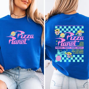 May include: A royal blue sweatshirt featuring the "Pizza Planet" logo in pink and yellow. The back of the sweatshirt has the "Pizza Planet" logo, alien characters, and text that reads "Friday Night Kids Night".