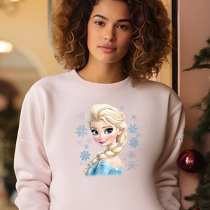 May include: A pink sweatshirt with a graphic print of Elsa from the movie Frozen. The graphic features Elsa's face and hair, with a blue and white dress and snowflakes surrounding her.