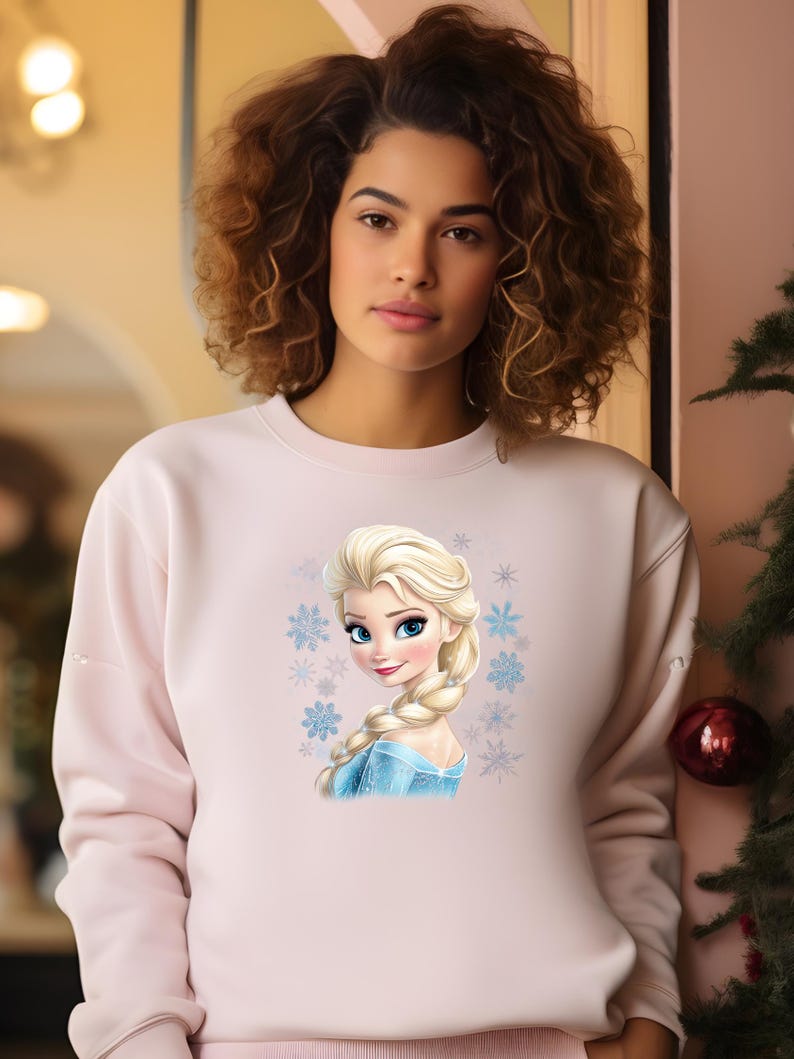 May include: A pink sweatshirt with a graphic print of Elsa from the movie Frozen. The graphic features Elsa's face and hair, with a blue and white dress and snowflakes surrounding her.