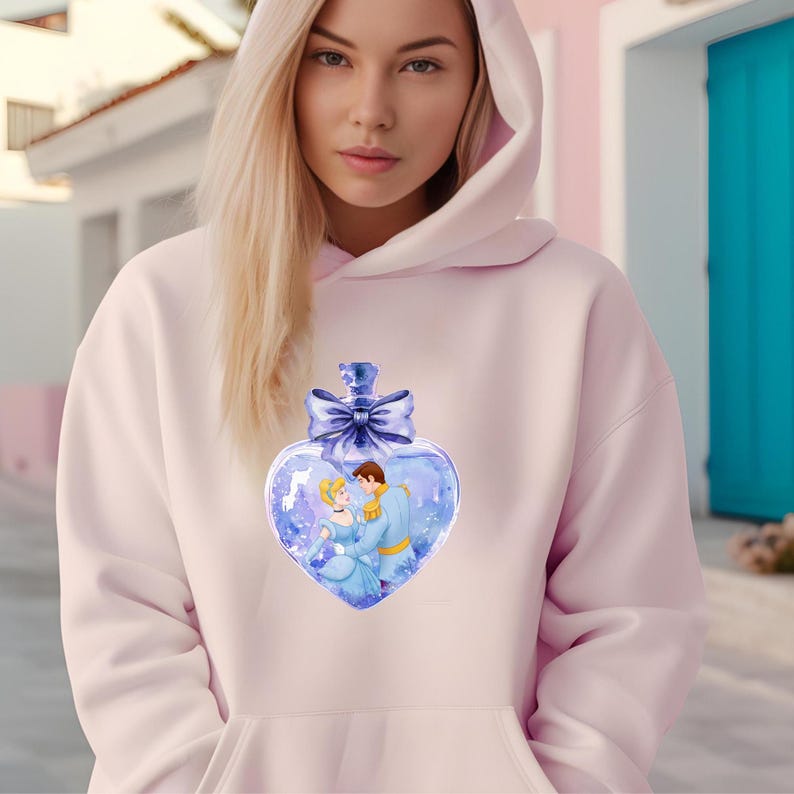 Puede incluir: Una sudadera con capucha rosa con un dise&ntilde;o de botella de vidrio en forma de coraz&oacute;n con Cenicienta y el Pr&iacute;ncipe Azul. La botella tiene un lazo azul y est&aacute; llena de una sustancia brillante azul y blanca.