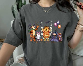 May include: Dark gray t-shirt featuring a Halloween-themed graphic of Winnie the Pooh and friends in costumes. Tigger is dressed as a witch, Pooh as a pumpkin, and Eeyore is reading a book. Balloons and ghosts complete the festive design.