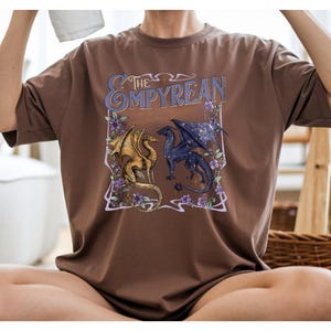 Pode incluir: Camiseta marrom com um design gr&aacute;fico. O design apresenta o texto "The Ompyrean" acima de dois drag&otilde;es. Um &eacute; dourado, o outro azul escuro com asas estreladas. Flores roxas e linhas decorativas emolduram os drag&otilde;es.