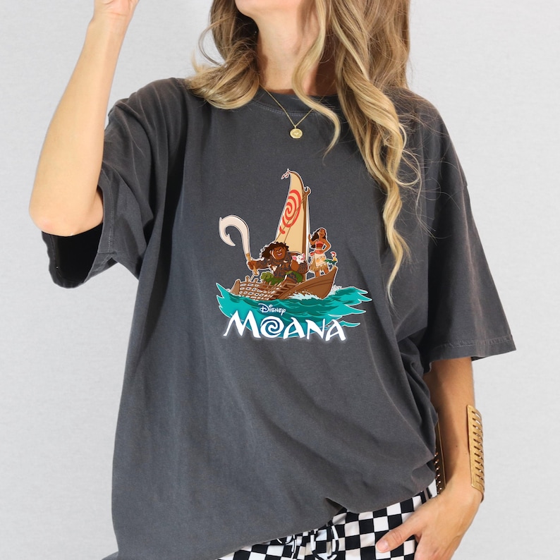 May include: A dark gray t-shirt with a graphic print of the Disney characters Moana and Maui sailing on a boat. The text "Disney Moana" is printed below the image.