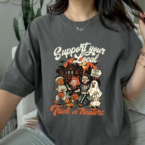 A person wearing a gray t-shirt with a Halloween-themed graphic design featuring a skeleton, ghost, and other spooky characters.