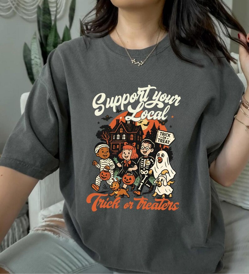A person wearing a gray t-shirt with a Halloween-themed graphic design featuring a skeleton, ghost, and other spooky characters.