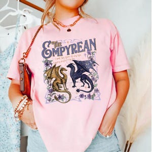 Pode incluir: T-shirt rosa claro com um design gr&aacute;fico com o texto "The Empyrean" e dois drag&otilde;es. Um &eacute; dourado, o outro azul escuro com asas estreladas. O design inclui detalhes florais roxos e a frase "I had the time of my life fighting dragons with you."