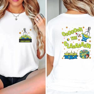 May include: White t-shirt featuring a cartoon design of green aliens and a claw machine. The front of the shirt has a small graphic with the text "EST.1995". The back of the shirt has the text "Ooooh the Claaaaaw" with more alien graphics.