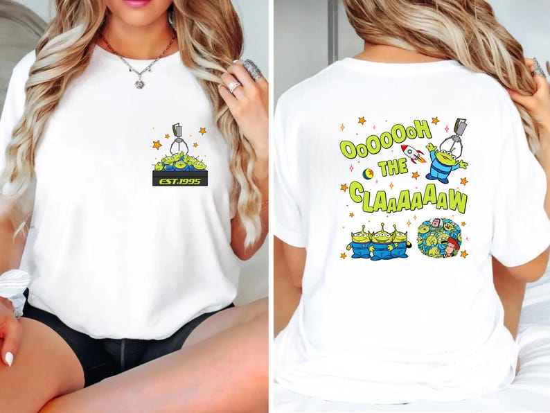 May include: White t-shirt featuring a cartoon design of green aliens and a claw machine. The front of the shirt has a small graphic with the text "EST.1995". The back of the shirt has the text "Ooooh the Claaaaaw" with more alien graphics.