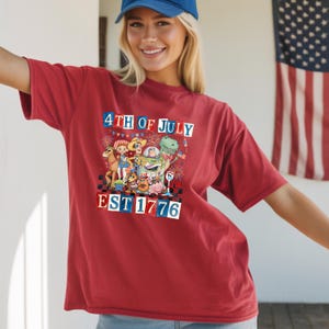 A smiling woman wearing a red t-shirt with a graphic design celebrating the 4th of July, including a dinosaur, fireworks, and the text "EST. 1776".