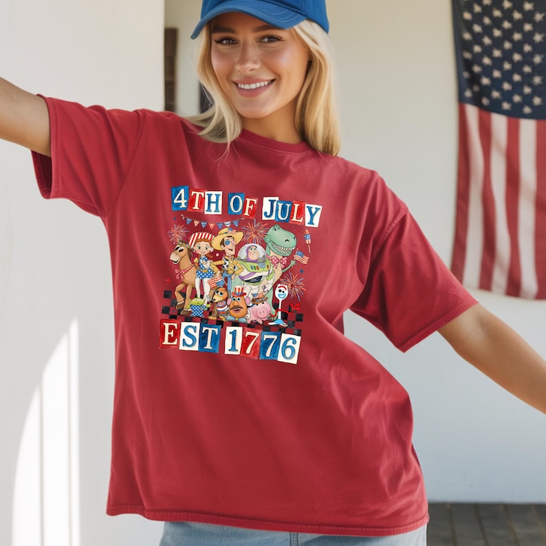 A smiling woman wearing a red t-shirt with a graphic design celebrating the 4th of July, including a dinosaur, fireworks, and the text "EST. 1776".