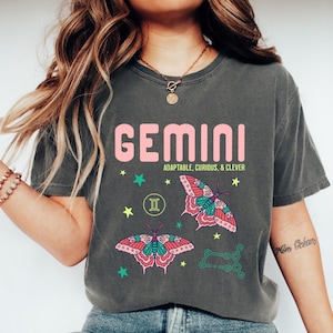Może przedstawiać: Ciemnoszara koszulka z napisem "GEMINI" w kolorze r&oacute;żowym i frazą "ADAPTABLE, CURIOUS, & CLEVER" poniżej. Koszulka ma grafiki motyli i gwiazd w kolorze r&oacute;żowym, zielonym i ż&oacute;łtym oraz symbol zodiaku Bliźnięta.