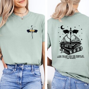A woman wearing a light green t-shirt with a graphic design on the back. The design features a dragon, a crescent moon, and a book, along with a quote that reads "I am the sky and the power of every storm I have ever been, I am infinite".