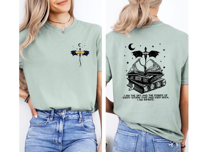 A woman wearing a light green t-shirt with a graphic design on the back. The design features a dragon, a crescent moon, and a book, along with a quote that reads "I am the sky and the power of every storm I have ever been, I am infinite".