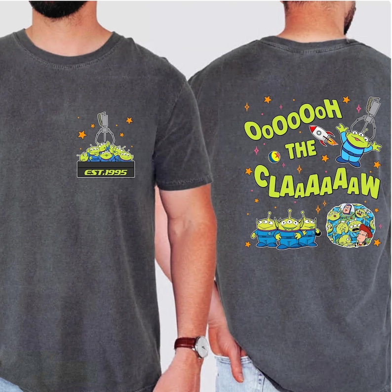 May include: Dark grey t-shirt featuring a cartoon claw machine graphic with green aliens and the text "EST.1995" on the front. The back of the shirt has the text "OooOooh THE CLAAAAAAW" with alien characters and a rocket ship.