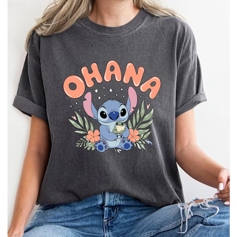 May include: Dark gray t-shirt featuring the word "OHANA" in orange bubble letters above a cartoon character. The character is blue with large ears, holding a small green creature, surrounded by flowers and leaves.