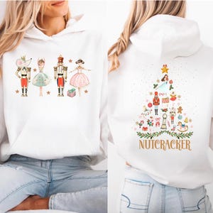 May include: White hooded sweatshirt featuring a Nutcracker theme. The front displays four characters: a mouse, a ballerina, a nutcracker soldier, and another ballerina. The back features a Christmas tree design with the word "NUTCRACKER" below.