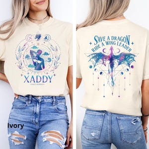 May include: A cream colored t-shirt with a graphic design of a dragon and the text "Save a Dragon Ride a Wing Leader". The back of the shirt has a graphic design of a person with the text "Xaddy - Xaddys Kingdom".