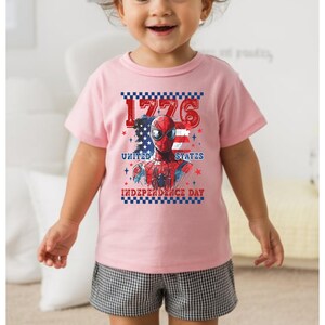May include: A light pink t-shirt featuring a graphic of Spider-Man with the American flag and the text "1776 United States Independence Day". The shirt is worn by a child.