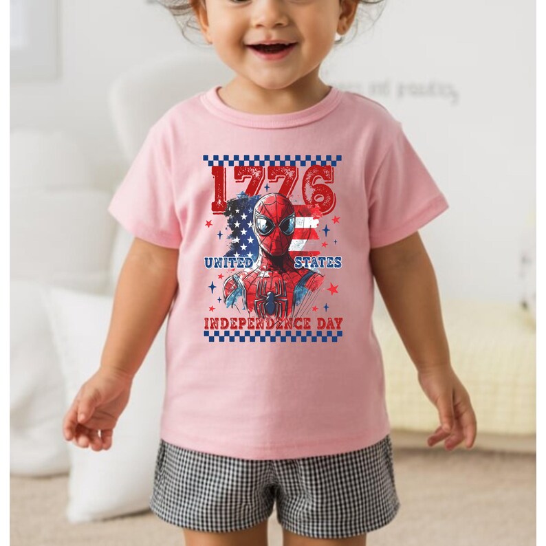 May include: A light pink t-shirt featuring a graphic of Spider-Man with the American flag and the text "1776 United States Independence Day". The shirt is worn by a child.