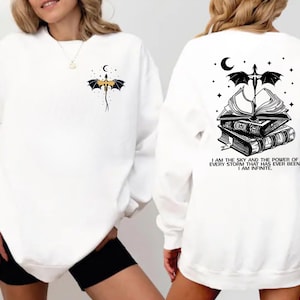 a woman wearing a white sweatshirt with a black and white graphic design on the back. The design includes a dragon, a crescent moon, and a book, along with a quote that reads "I AM THE SKY AND THE POWER OF EVERY STRAIGHT CURVE HAS BEEN ININITIATIVE".