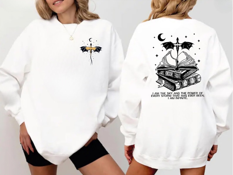 a woman wearing a white sweatshirt with a black and white graphic design on the back. The design includes a dragon, a crescent moon, and a book, along with a quote that reads "I AM THE SKY AND THE POWER OF EVERY STRAIGHT CURVE HAS BEEN ININITIATIVE".