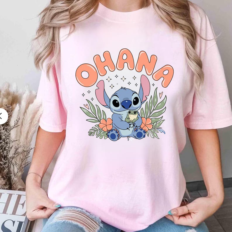 May include: Pink t-shirt featuring a cartoon illustration of Stitch from Lilo & Stitch. The blue character is holding a small frog, surrounded by flowers and leaves. The word "OHANA" is printed above the character in orange bubble letters.