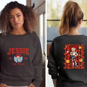 May include: Dark gray sweatshirt featuring the name "JESSIE" in red block letters with a cartoon image of Jessie from Toy Story. The back of the sweatshirt displays a larger graphic of Jessie with the name "JESSIE" in a retro design.