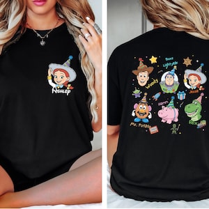Puede incluir: Camiseta negra con un dibujo animado de Jessie de Toy Story en la parte delantera, con el nombre "Ashley" debajo. La parte trasera de la camiseta muestra varios personajes de Toy Story, incluyendo a Woody, Buzz Lightyear y Mr. Potato Head.